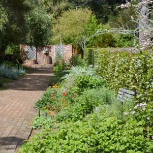 A serene brick pathway winds through a lush garden filled with vibrant flowers and diverse greenery. The path leads to a brick wall with two lattice windows and a central fountain, surrounded by mature trees and hedges. Benches are placed along the path, inviting visitors to sit and enjoy the tranquil setting.
