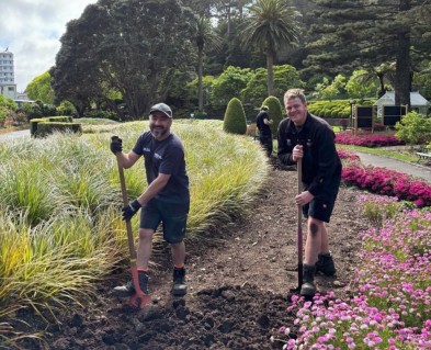 Two uniformed men with spades turning over the soil on a garden path. Flowers and grasses are on either side with a view of the more paths and garden beds behind them. 