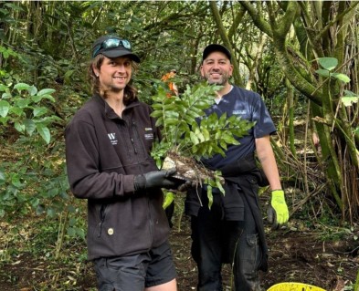 two men smiling while holding up a shoe with a fern growing out of it. They are in a New Zealand Native forest.